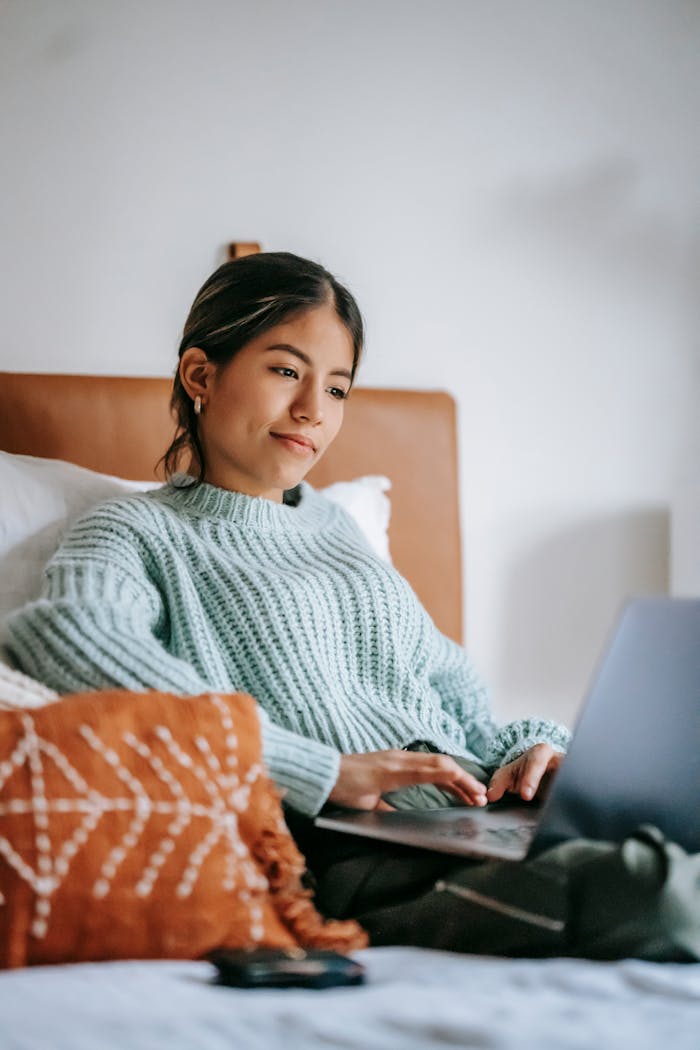 Happy young ethnic female using netbook while searching important information on comfortable bed with pillows